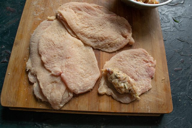 Раскладываем запанированные куриные отбивные на рабочей поверхности. На один край каждого мясного пласта выкладываем примерно 50 граммов начинки.