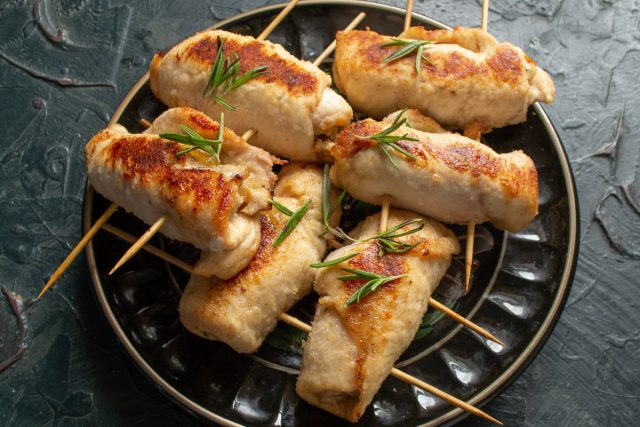Выкладываем готовые куриные рулетики на сервировочное блюдо. Посыпаем горячую закуску свежими листиками розмарина и незамедлительно подаем к столу.