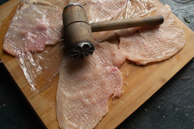 Выкладываем ломтики куриного филе на разделочную доску и накрываем пищевой пленкой. Отбиваем мясо кулинарным молотком с обеих сторон до тонкости.