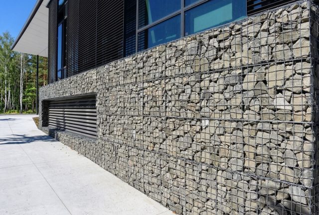 Габионы используют для оформления фасадов, создавая эффект массивной каменной поверхности. Такая отделка улучшает звукоизоляцию и придает зданию современный, архитектурный характер.
