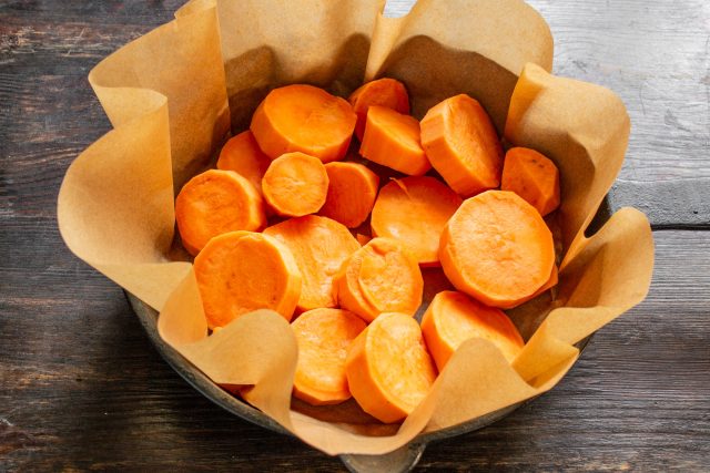 Застилаем чугунную сковороду или огнеупорную форму листом пергамента для выпечки. Выкладываем нарезанные кружочки батата в емкость, распределяя их по дну. Застилаем чугунную сковороду или огнеупорную форму листом пергамента для выпечки. Выкладываем нарезанные кружочки батата в емкость, распределяя их по дну.