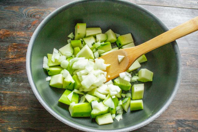 Очищаем свежий репчатый лук от шелухи, мелко шинкуем его ровными кубиками на разделочной доске и аккуратно добавляем в общую кастрюлю к нарезанным кабачкам.