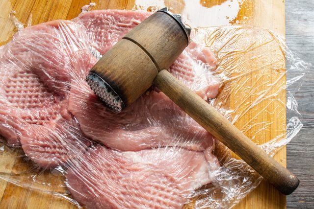 Выкладываем нарезанное мясо на разделочную доску и накрываем пищевой пленкой. Тщательно отбиваем молоточком с двух сторон, чтобы защитить кухню от мясных брызг в процессе.