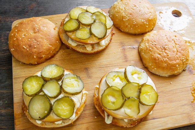 Накрываем горячую куриную котлету тонким ломтиком копченого сыра для быстрого подтаивания, нарезаем маринованный или квашеный огурец тонкими кружочками и распределяем их поверх сыра.